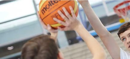 Spieler wirft einen Basketball auf einen Basketballkorb, ein Verteidiger versucht zu blocken