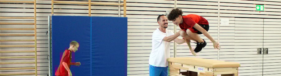 Kinder im Sportunterricht springen mit Hilfestellung über einen Sprungkasten