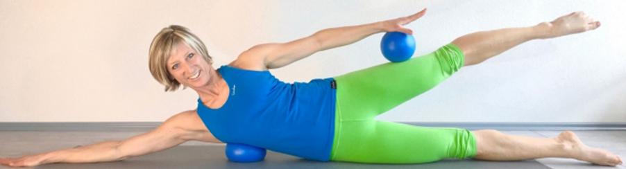Frau führt in Seitenlage auf einer Gymnastikmatte Pilatesübungen mit zwei kleinen blauen Pilatesbällen aus