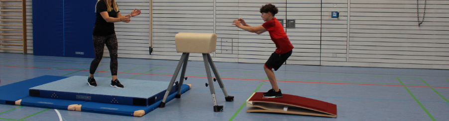 Schüler springt vom Sprungbrett über den Turnbock mit Fallschutzmatten und Lehrkraft
